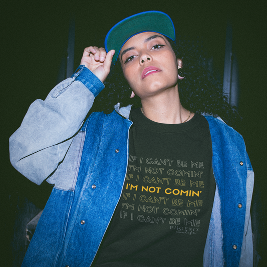 Woman with long curly hair, a cap, and a sweatshirt on under a denim jacket. The sweatshirt says If I Can't Be Me I'm Not Comin'. Sweatshirt is black with yellow writing.